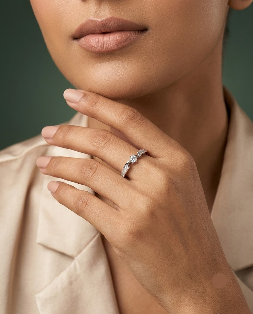 A close-up lifestyle shot of a model wearing a delicate silver band ring with a small clear heart-shaped gemstone on her finger, paired with a beige satin blazer against a dark green background.
