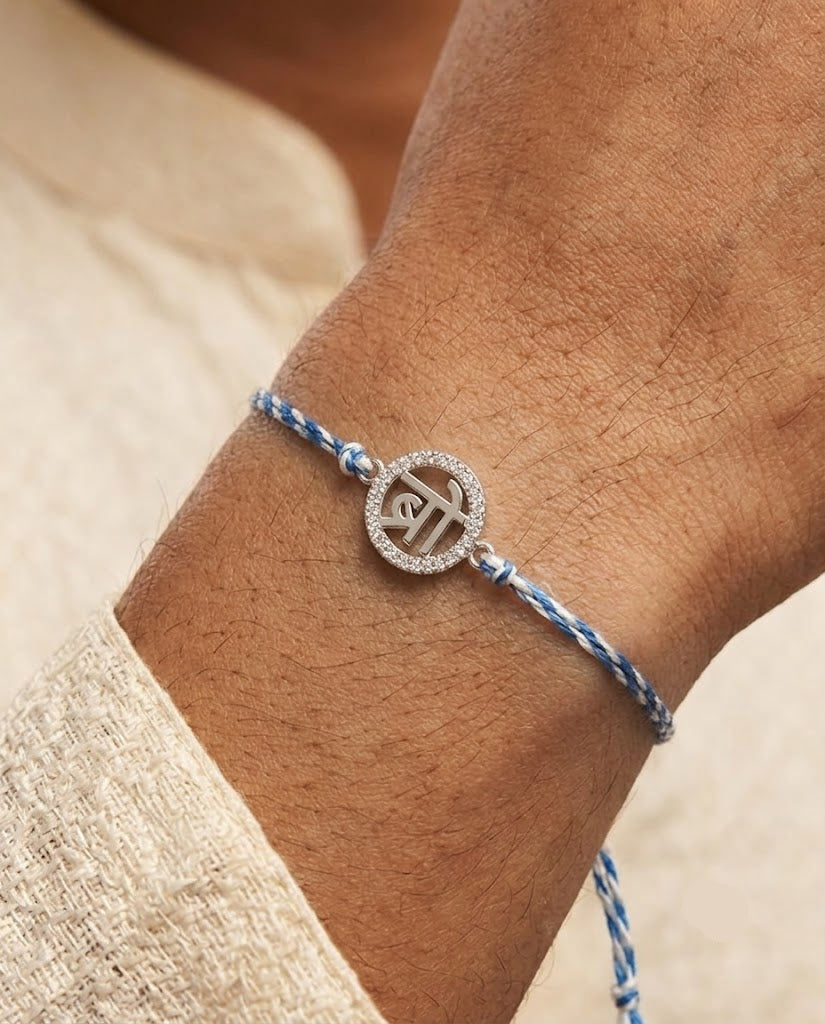 A close-up of a person's wrist wearing a delicate braided blue and white string bracelet with a silver-toned circular pendant featuring a Devanagari script symbol.