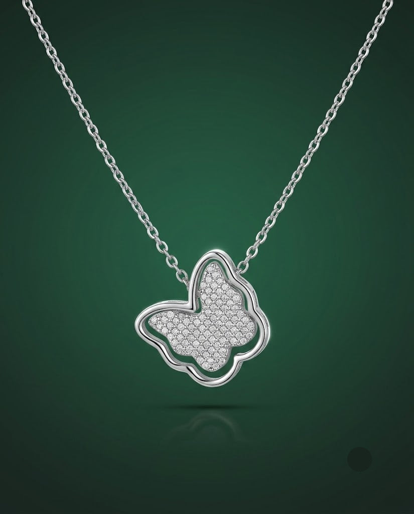 A silver butterfly pendant necklace with a textured pavé center, suspended from a delicate silver chain against a dark green gradient background.