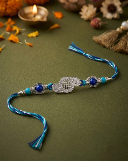 A traditional Indian Rakhi with a silver-toned central ornament, blue beads, turquoise accents, and blue tassels, displayed on a textured olive green surface with festive decorative props like a lit diya, dried flowers, and marigold petals.