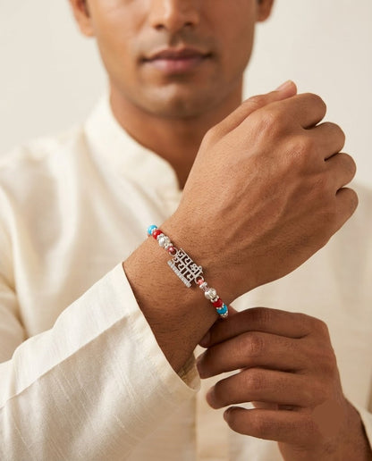A man wearing a silver-toned metal bracelet with colorful red, blue, and white beads on his wrist, wearing a white shirt against a warm beige background.