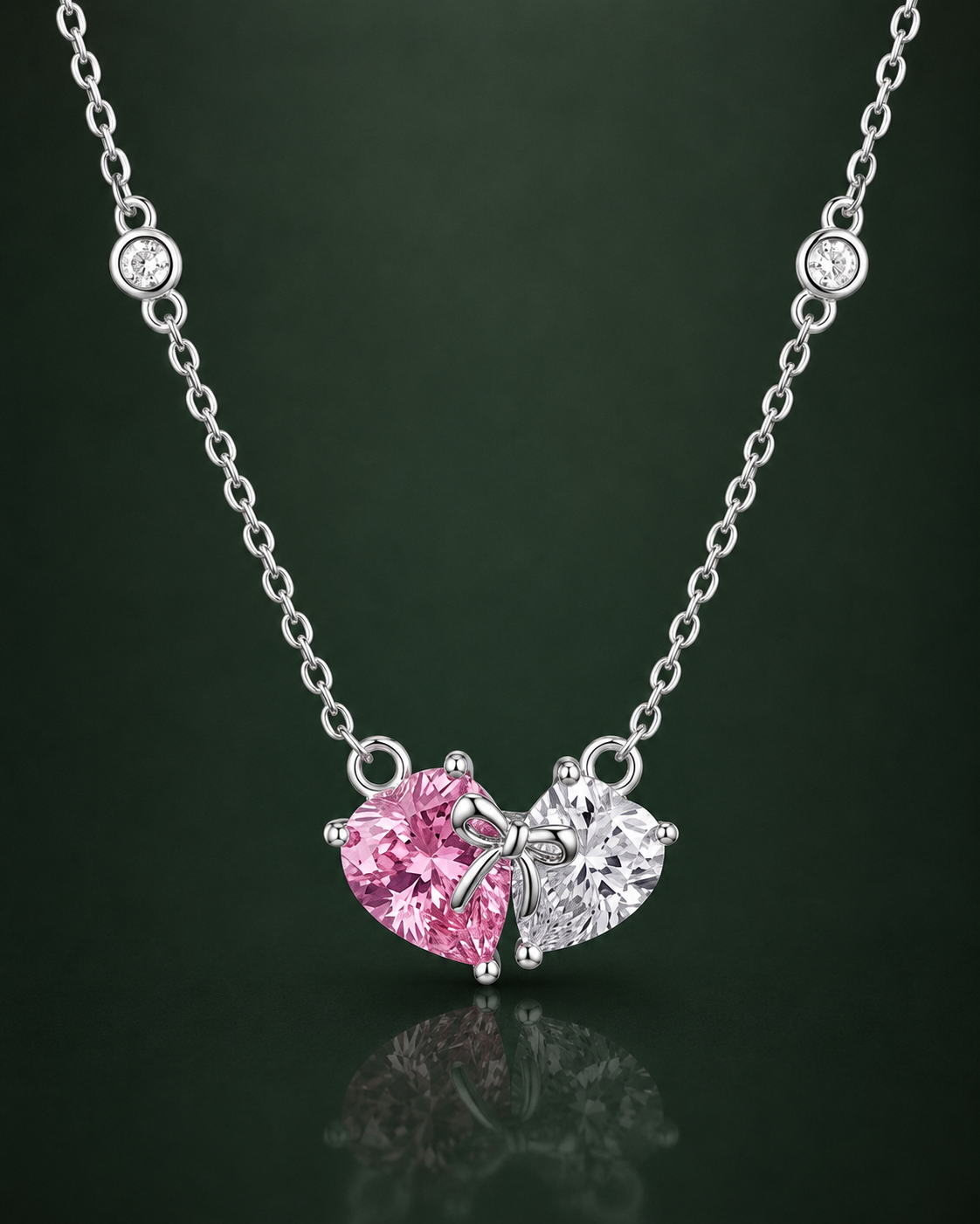 A silver necklace with a heart-shaped pendant featuring one pink gemstone and one clear gemstone, displayed against a dark green reflective background.