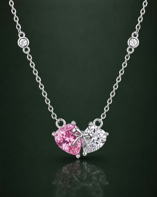 A silver necklace with a heart-shaped pendant featuring one pink gemstone and one clear gemstone, displayed against a dark green reflective background.
