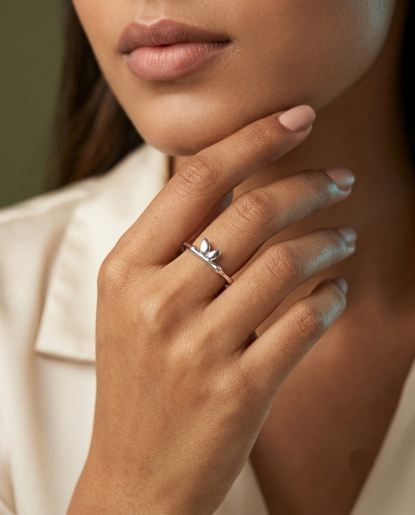 A close-up lifestyle shot of a model wearing a delicate silver ring with a small leaf motif on her finger, paired with a cream-colored collared shirt.