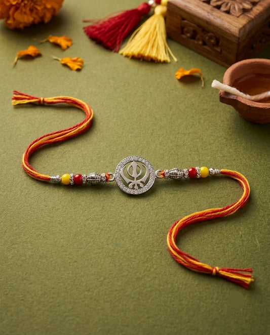 A Sikh Rakhi with a silver Khanda emblem, accented with red and yellow beads, displayed on an olive green textured surface with festive decorative props.