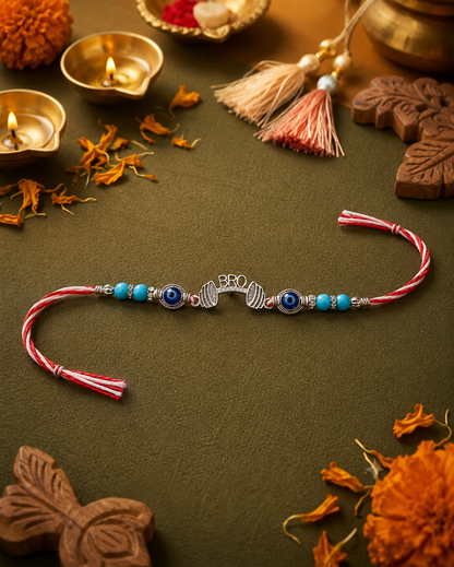 A silver-toned Hindu Rakhi beaded wristband with blue evil eye beads and red-and-white twisted tassel ends, displayed on a dark olive green surface surrounded by festive props including lit diya lamps, marigold flowers, and carved wooden ornaments.