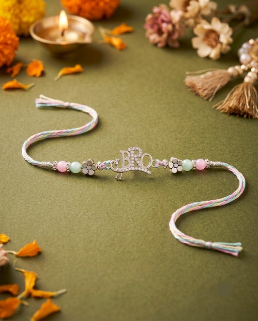 A silver-toned Rakhi bracelet with a central 'Rakhi' charm, decorated with pastel pink, mint green, and white beads, displayed on an olive green surface with decorative marigold petals, a lit diya, and dried flowers.