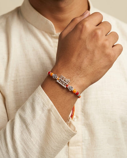 A close-up of a person wearing a red and orange beaded bracelet with a silver-toned 'Jai Ho' charm, paired with a cream-colored linen shirt.