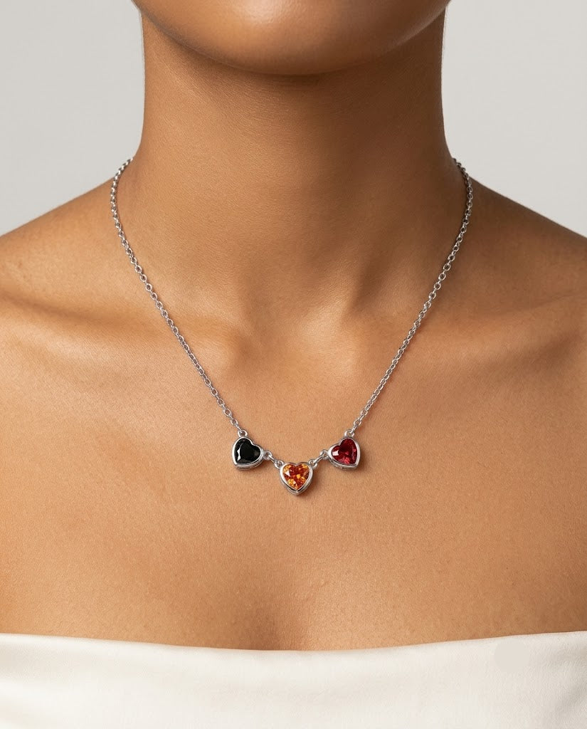 A silver chain necklace with three small heart-shaped gemstones in black, orange, and red, worn on a model's neck.