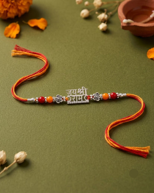 A traditional Indian Rakhi with a silver  central charm featuring Devanagari text, accented with red and orange beads, displayed on an olive green background with festive decorative props.