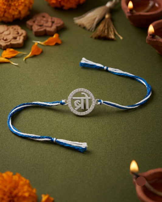 A blue and white braided Rakhi bracelet with a silver-toned circular pendant featuring the Devanagari script for 'Bro' (बro), displayed on a dark olive green background with festive decorative props like marigold flowers, lit diya lamps, and traditional stamps.