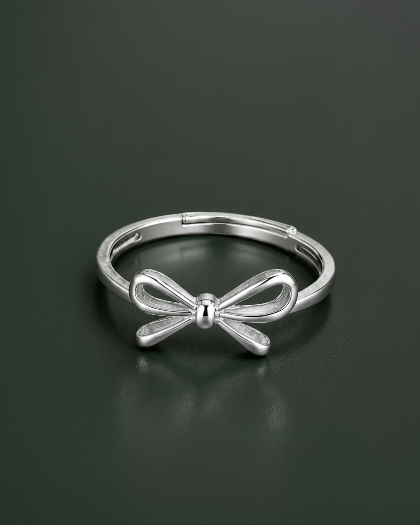 A polished silver metal ring featuring a bow-shaped centerpiece, displayed on a dark, reflective surface.