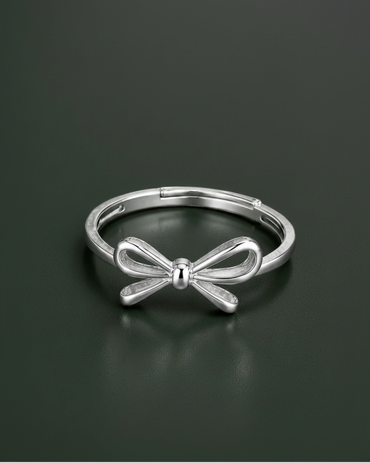 A polished silver metal ring featuring a bow-shaped centerpiece, displayed on a dark, reflective surface.
