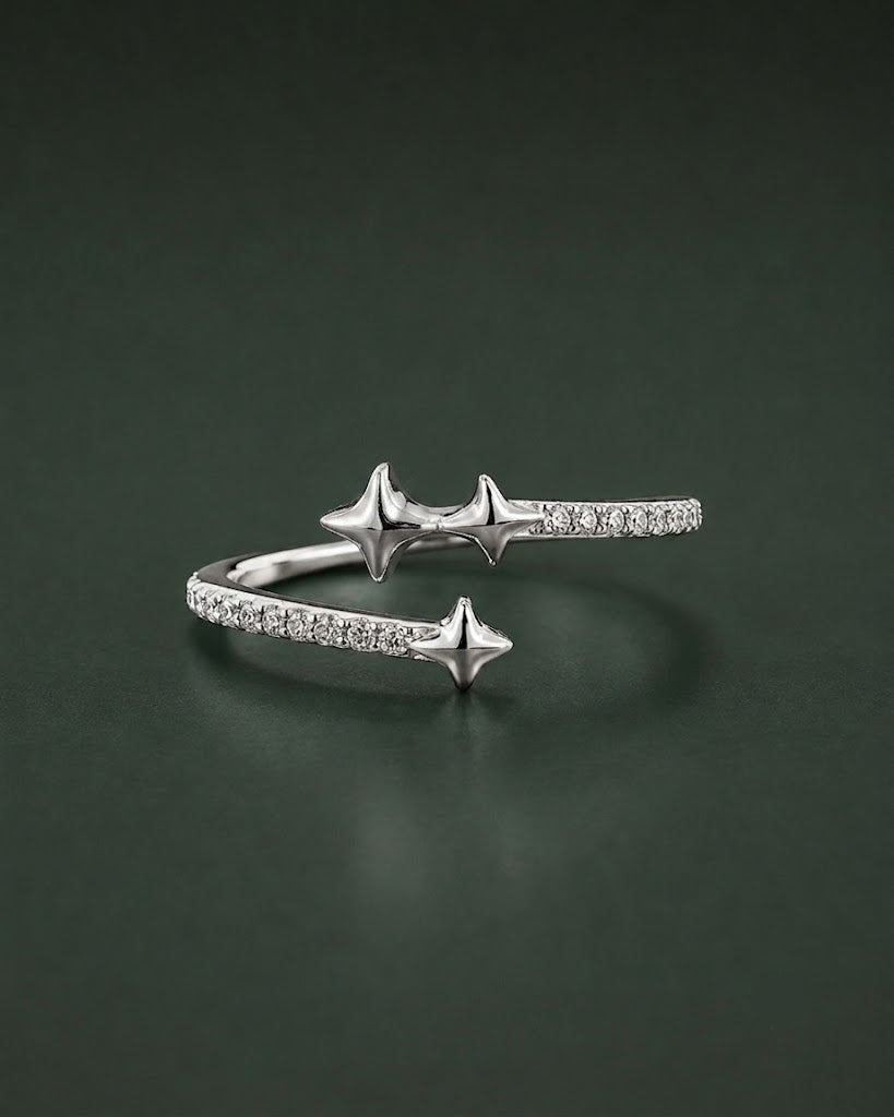 A silver open cuff ring featuring three star-shaped charms and pavé-set clear stones, displayed on a dark green reflective surface.