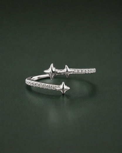 A silver open cuff ring featuring three star-shaped charms and pavé-set clear stones, displayed on a dark green reflective surface.
