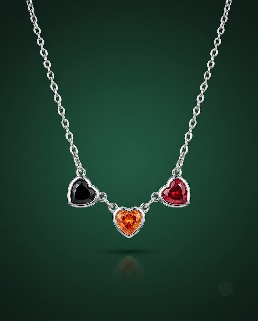 A silver heart pendant necklace featuring three dangling heart charms in black, red, and orange, suspended from a silver chain against a dark green gradient background.
