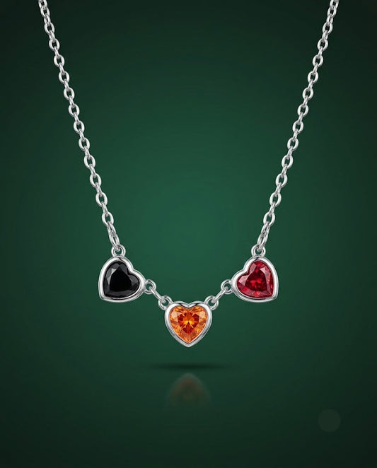 A silver heart pendant necklace featuring three dangling heart charms in black, red, and orange, suspended from a silver chain against a dark green gradient background.