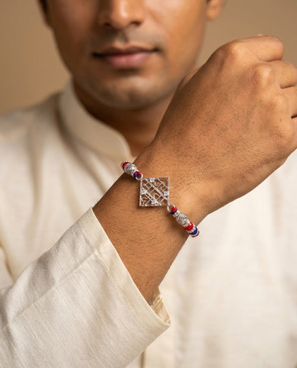 A close-up of a man wearing a silver-toned diamond-embellished bracelet with red and blue beaded accents on his wrist, wearing a cream-colored shirt.