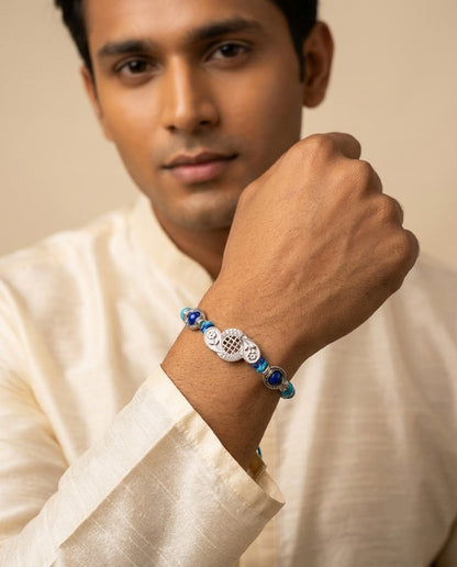 A man wearing a white kurta and holding his hand near his wrist, showcasing a beaded bracelet with white, blue, and silver elements.