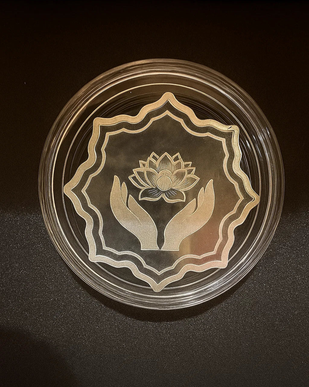 Decorative glass plate with engraved design of hands holding a lotus flower on a dark background