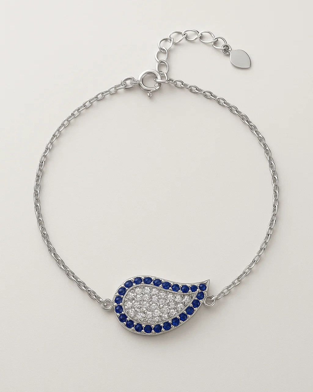 Silver bracelet with a teardrop-shaped charm featuring blue and clear stones on a light gray background.