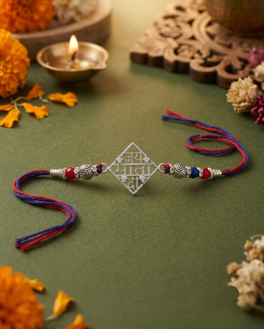 A silver-toned Rakhi with a central diamond-shaped charm featuring Devanagari script, adorned with red and blue thread tassels and metallic beads, displayed on a green surface with festive decorative props.
