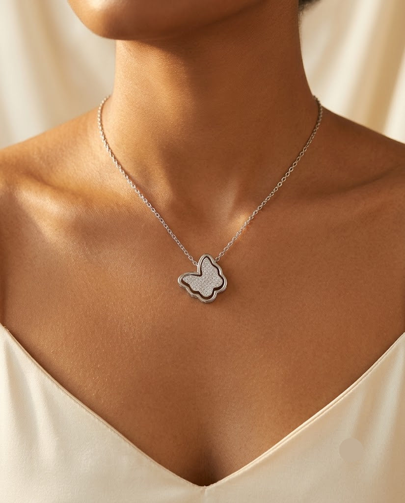 A close-up of a model wearing a delicate silver butterfly pendant necklace with a textured center, styled with a cream-colored satin top.