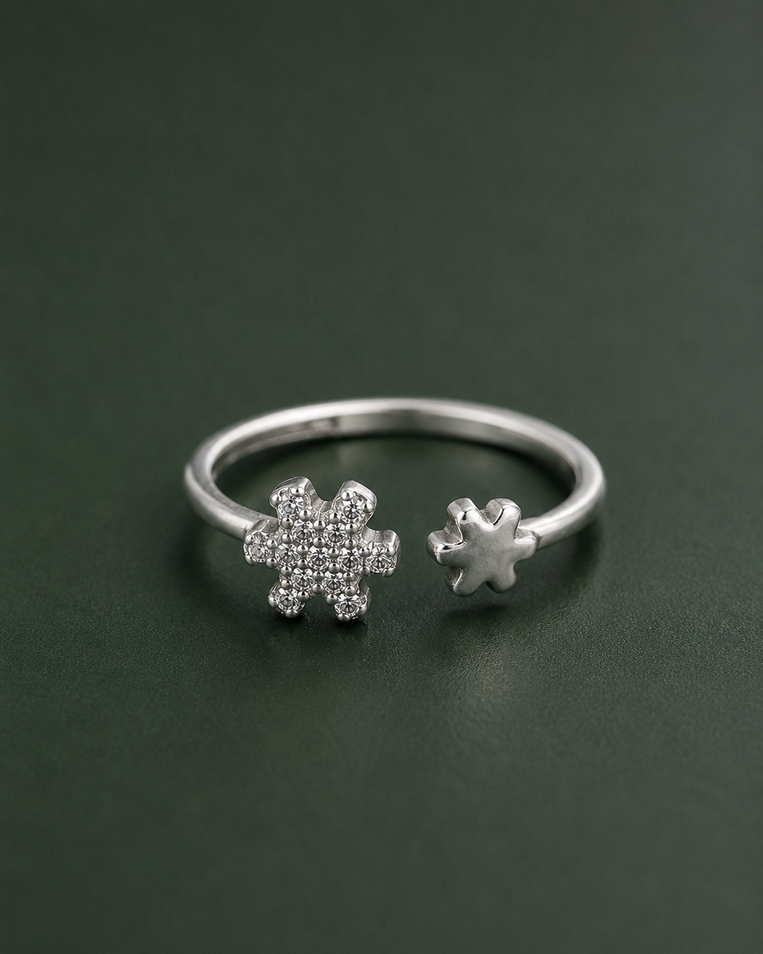 A silver open ring featuring two snowflake-shaped settings, one pavé-set with small clear stones and one plain, displayed on a dark green background.