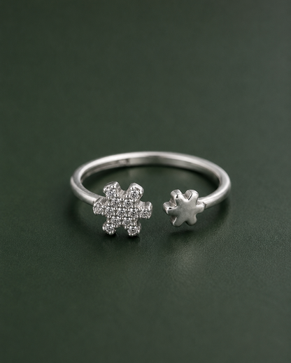 A silver open ring featuring two snowflake-shaped settings, one pavé-set with small clear stones and one plain, displayed on a dark green background.