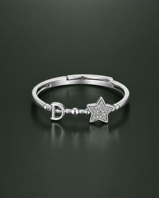 A silver- ring featuring a small 'D' charm and a star-shaped charm paved with clear stones, displayed on a dark reflective surface.