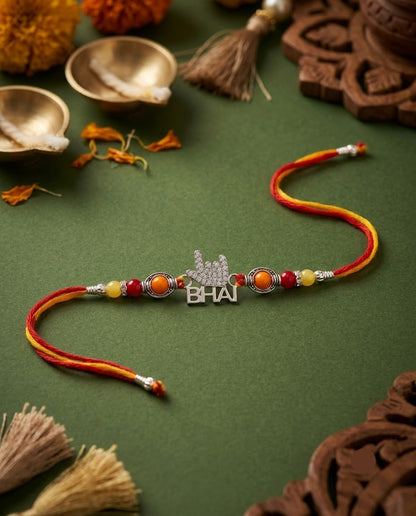 A traditional Indian Rakhi with a silver 'BHAI' nameplate and red, orange, and yellow beads, displayed on a dark green textured background with festive decorative props.