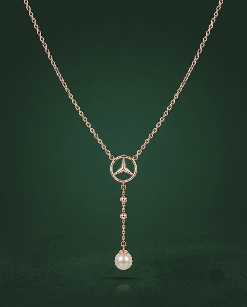 A rose gold necklace featuring a Mercedes-Benz three-pointed star logo pendant, with a dangling pearl bead at the bottom, set against a dark green background.