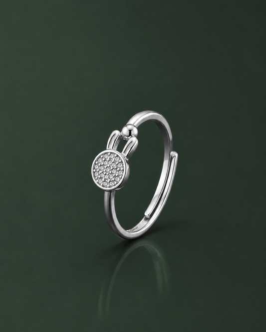 A silver open ring with a round pavé-set stone centerpiece and a small spherical accent above it, photographed on a dark green reflective surface.